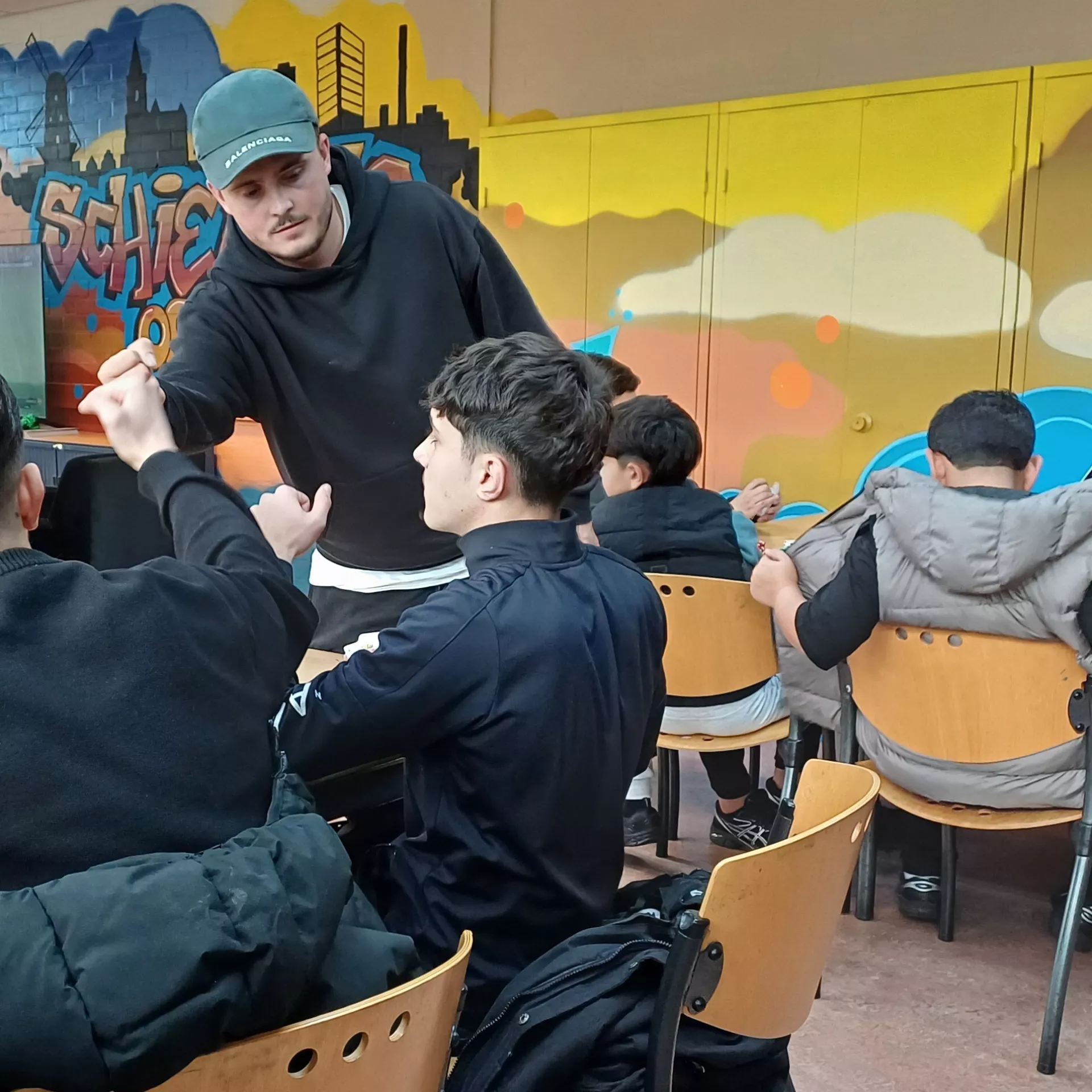 Kinderen zitten in een klaslokaal met kleurrijke muurschilderingen op de achtergrond.